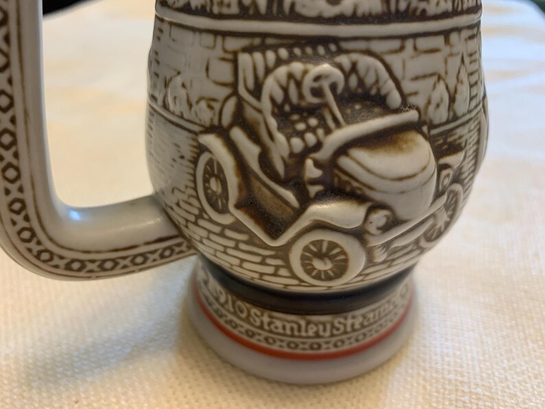 May include: A ceramic beer stein featuring a raised relief design of a vintage automobile on a brick-patterned background. The base of the stein includes the text 1910 Stanley Steamer, with a handle decorated in a geometric pattern and brown accents.