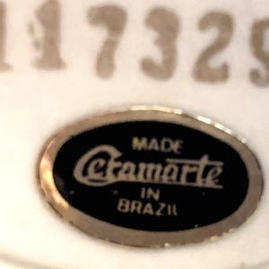 May include: An oval-shaped black label with a gold-toned border featuring the text "MADE Cramarte IN BRAZIL" in white lettering. Above the label, the number "117329" is printed in a large, light-colored font on a white background.