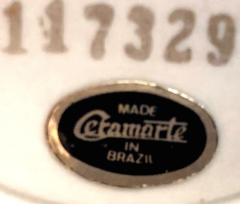 May include: An oval-shaped black label with a gold-toned border featuring the text "MADE Cramarte IN BRAZIL" in white lettering. Above the label, the number "117329" is printed in a large, light-colored font on a white background.