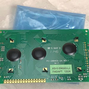 May include: A green circuit board with three black circular buttons and gold connectors. The board has various electronic components and text, including "PC-2004E6-2A REV.1" and "ASI-G-204AAS-LJ-FWD/W*T 12024". A blue plastic bag is in the background.