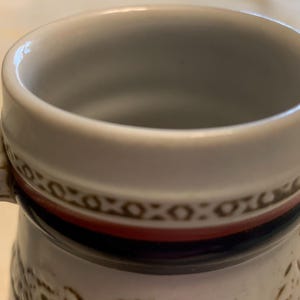 May include: A ceramic mug featuring a white interior and a thick, rounded rim. The exterior displays a decorative band with a brown geometric pattern, a solid red stripe, and a dark blue stripe. A handle with matching brown geometric detailing is attached.