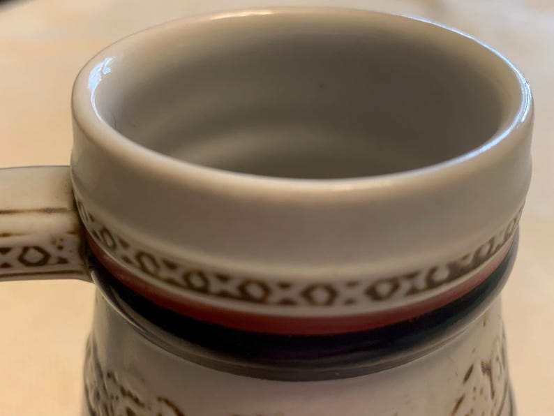May include: A ceramic mug featuring a white interior and a thick, rounded rim. The exterior displays a decorative band with a brown geometric pattern, a solid red stripe, and a dark blue stripe. A handle with matching brown geometric detailing is attached.