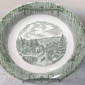 May include: A white ceramic plate with a green rural landscape design. The center features a detailed illustration of a village scene with houses, trees, and a horse-drawn carriage. The rim has a textured green pattern.