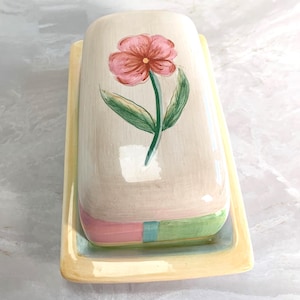 May include: A rectangular ceramic butter dish with a lid. The lid is painted with a pink flower and green leaves. The base is yellow, and the dish has a pink and green band around the middle.