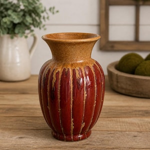 May include: A decorative red vase with vertical grooves and a brown rim. The vase sits on a wooden surface, showcasing its rustic charm. The vase is a stylish home decor item.