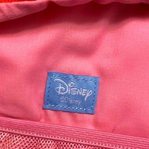 May include: A bright pink fabric interior featuring a rectangular blue woven label stitched in the center. The label displays the white Disney logo and the text Disney below it. A pink mesh pocket with a zipper is visible along the bottom edge.