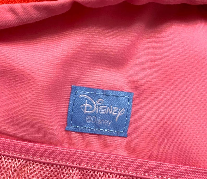 May include: A bright pink fabric interior featuring a rectangular blue woven label stitched in the center. The label displays the white Disney logo and the text Disney below it. A pink mesh pocket with a zipper is visible along the bottom edge.