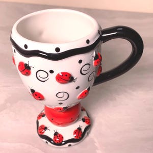 May include: A white ceramic coffee mug with a black handle and a red base. The mug is decorated with black polka dots, black curved lines, and red ladybug accents. The mug is designed with a whimsical, playful theme.