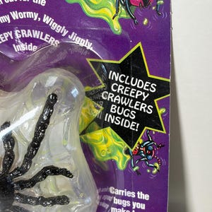 May include: A purple package with a clear plastic bubble containing a black spider toy. The package has green and yellow accents, with text that reads "Includes Creepy Crawlers Bugs Inside!" and "Creepy Crawlers Inside".
