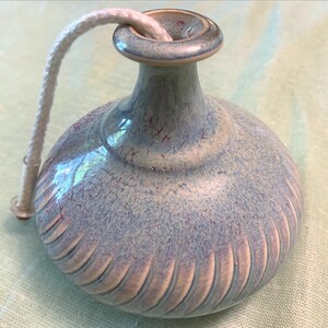 May include: A ceramic oil lamp with a light blue and lavender glaze. The lamp has a round body with vertical grooves and a slender neck. A white wick extends from the top. The lamp is designed for aromatherapy or ambiance.