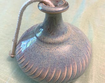 May include: A ceramic oil lamp with a light blue and lavender glaze. The lamp has a round body with vertical grooves and a slender neck. A white wick extends from the top. The lamp is designed for aromatherapy or ambiance.