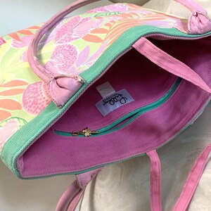 May include: A colorful tote bag with a floral pattern in shades of pink, orange, and green. The bag has pink handles and a green trim. The interior is pink with a teal zipper and a gold zipper pull.