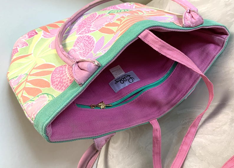May include: A colorful tote bag with a floral pattern in shades of pink, orange, and green. The bag has pink handles and a green trim. The interior is pink with a teal zipper and a gold zipper pull.