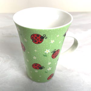May include: A tall, light green ceramic mug with a white interior and handle. The mug is decorated with red ladybugs and small white flowers. The mug has a slightly flared shape and is suitable for hot or cold beverages.