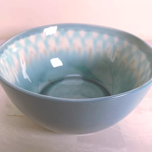 May include: A ceramic bowl with a light blue exterior and a glossy, teal interior. The interior features a white and tan pattern near the rim. The bowl has a smooth, rounded shape and is suitable for serving food.