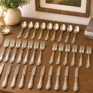 May include: A collection of antique silverware, including forks, spoons, knives, and a ladle, arranged on a wooden surface. The silverware has ornate, detailed handles and a shiny, reflective finish. A decorative vase with greenery and framed artwork are in the background.