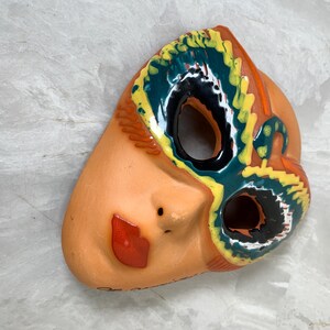 May include: A ceramic mask with a peach-colored face, a red lip, and a black dot for a nostril. The eye and forehead area are decorated with yellow, teal, orange, and black patterns. The mask has two eye holes.