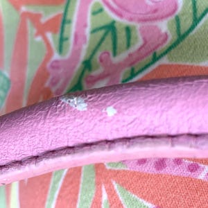 May include: Close-up of a pink leather handle with white spots. The handle is set against a colorful background with a pattern of pink flamingos, green leaves, and orange and white shapes.