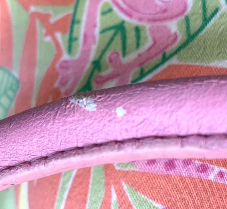 May include: Close-up of a pink leather handle with white spots. The handle is set against a colorful background with a pattern of pink flamingos, green leaves, and orange and white shapes.