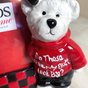 May include: A ceramic bear figurine with a white head, black bow, and red sweater. The sweater has the text "Do These Make My Butt Look By?" The bear wears black pants and white shoes. The figurine is part of a decorative frame.