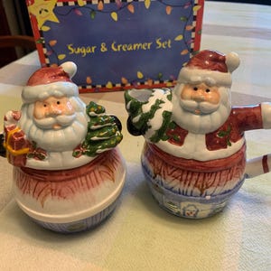 May include: A ceramic sugar and creamer set featuring Santa Claus figures. The sugar bowl has a lid with Santa holding a gift and tree. The creamer is shaped like Santa with a candy cane handle. The box is labeled Sugar & Creamer Set.