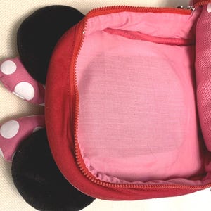 May include: A red backpack with a pink interior lining and a zipper closure. The exterior features two black circular ears and two pink fabric appendages with white polka dots extending from the side, resembling a cartoon character design.