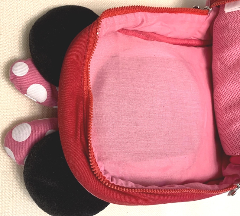 May include: A red backpack with a pink interior lining and a zipper closure. The exterior features two black circular ears and two pink fabric appendages with white polka dots extending from the side, resembling a cartoon character design.