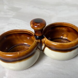 May include: A double-bowl ceramic condiment server featuring a central handle. The piece has a glossy finish with a two-tone color scheme, displaying a dark brown glaze on the upper rims and handle, transitioning into a creamy off-white base.