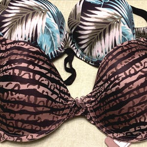 May include: Two bras with different patterns. One has a brown and black zebra stripe pattern. The other has a tropical leaf print in shades of blue, white, and brown. Both bras have underwire support and adjustable straps.