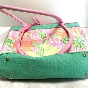 May include: A pink and green tote bag with a tropical floral pattern. The bag features pink handles and trim, a green base, and gold-colored rivets. The bag's design includes pink frogs, leaves, and flowers.