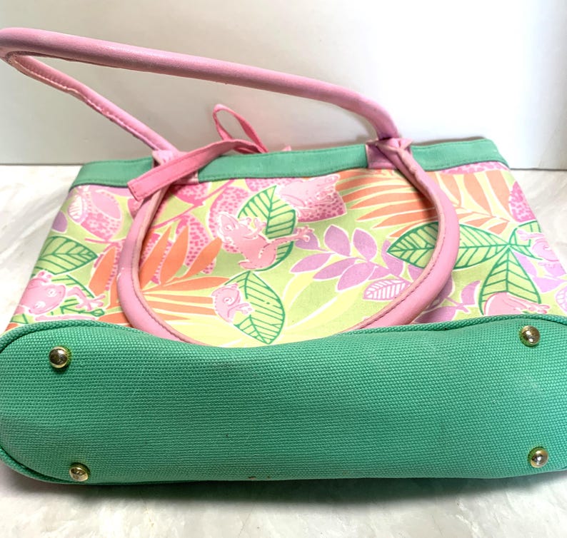 May include: A pink and green tote bag with a tropical floral pattern. The bag features pink handles and trim, a green base, and gold-colored rivets. The bag's design includes pink frogs, leaves, and flowers.