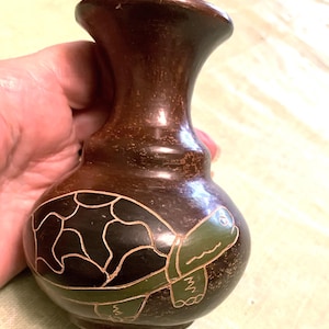 May include: A small, brown ceramic vase with a turtle design. The turtle is depicted in black and green, with white outlines. The vase has a rounded body and a flared neck. The vase is held in a hand.