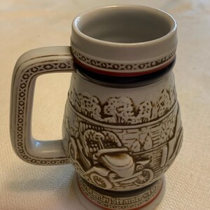 May include: A ceramic beer stein featuring a raised relief design of a vintage Stanley Steamer automobile. The mug has a light gray body with brown detailing, a sturdy handle, and a red and black band near the rim and base with text at the bottom.