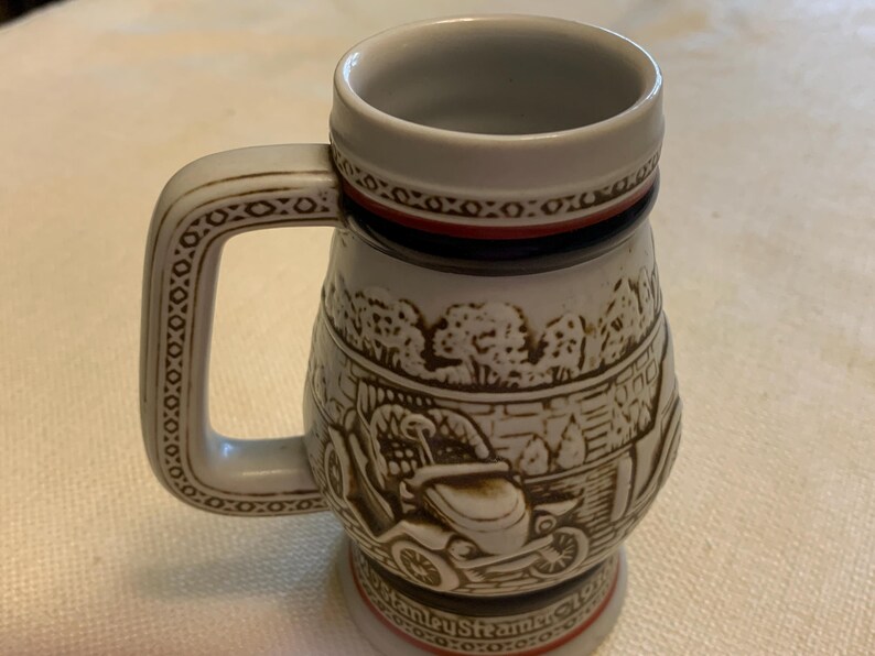 May include: A ceramic beer stein featuring a raised relief design of a vintage Stanley Steamer automobile. The mug has a light gray body with brown detailing, a sturdy handle, and a red and black band near the rim and base with text at the bottom.
