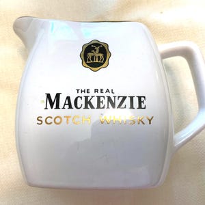 May include: A white ceramic pitcher featuring a black and gold logo of two deer inside a scalloped circle. The text reads THE REAL MACKENZIE in black and SCOTCH WHISKY in gold lettering. The pitcher has a curved handle and a pouring spout.
