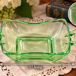May include: A light green glass dish with two side handles, measuring 3.5 inches wide and 5 inches long. The glass features a molded Heisey mark of a diamond with an H inside, visible in an inset circle on the upper right side of the image.