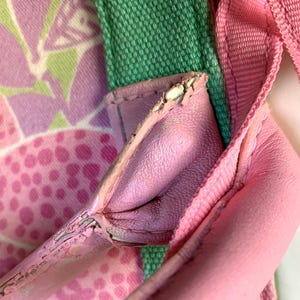 May include: Close-up of a pink handbag with a floral pattern. The bag features a pink leather strap and a green fabric strap. The floral pattern includes pink and white shapes. The bag's leather shows signs of wear.