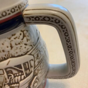 May include: A ceramic beer stein featuring a white base with brown embossed relief illustrations of a horse-drawn carriage. The handle is decorated with a geometric pattern in brown, and the rim is accented with a thin red band and a brown border.