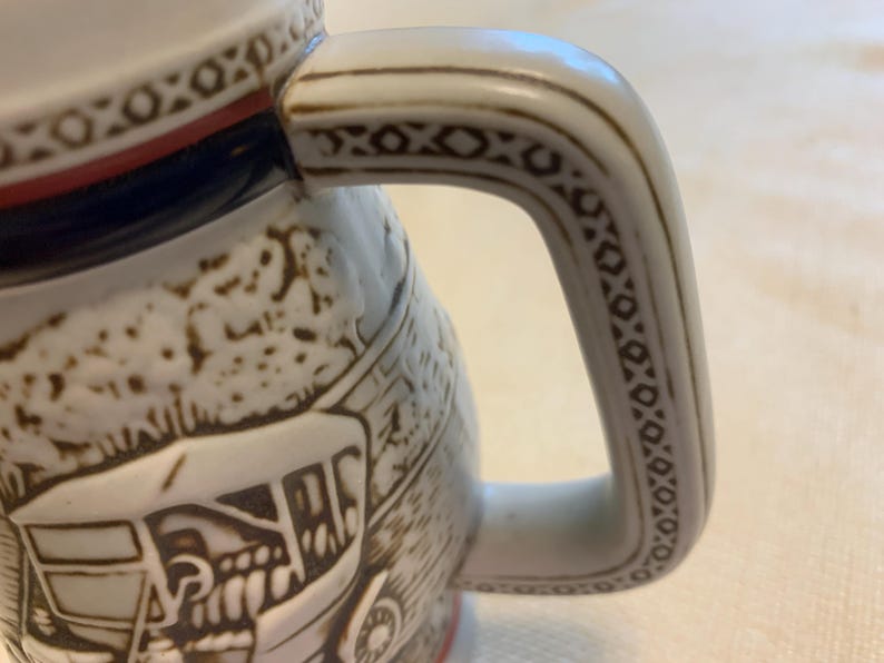 May include: A ceramic beer stein featuring a white base with brown embossed relief illustrations of a horse-drawn carriage. The handle is decorated with a geometric pattern in brown, and the rim is accented with a thin red band and a brown border.