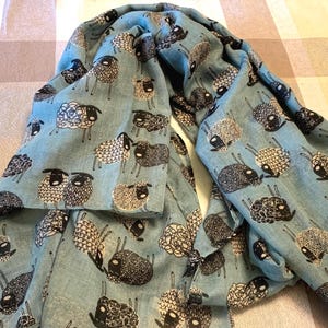 May include: A light blue scarf featuring a repeating pattern of black and white sheep illustrations. The sheep are depicted in various orientations with textured wool patterns, creating a consistent graphic design across the entire fabric surface.