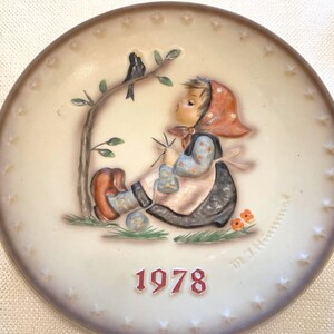 May include: A round ceramic collector plate featuring a raised relief illustration of a child wearing a red headscarf and blue patterned clothing, knitting while sitting under a tree with a black bird. The year 1978 is printed in red at the bottom.