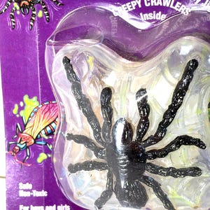 May include: A black plastic spider toy inside a clear plastic blister pack. The packaging is purple with cartoon illustrations of insects and the text "CREEPY CRAWLERS Inside". The packaging also states "Safe Non-Toxic" and "For boys and girls ages 8 & up."