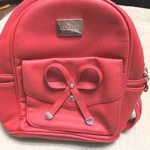 Puede incluir: Una mochila roja con un diseño en forma de lazo en el bolsillo delantero. La mochila tiene una pequeña placa metálica rectangular con la palabra "LCFUN". La bolsa tiene una cadena en el lateral.