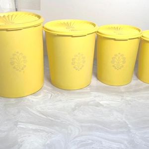 May include: Four yellow plastic canisters with lids. Each canister has a floral design and varies in size. The canisters are arranged from largest to smallest. The lids have a textured design.