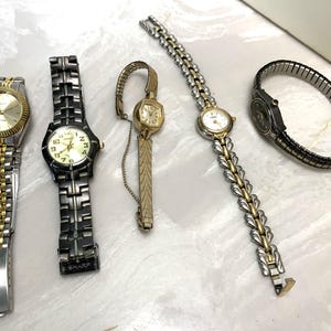 May include: A collection of five wristwatches with various designs. The watches feature gold and silver tones, with different band styles including link, stretch, and chain. The watch faces are round and display numbers or markers.