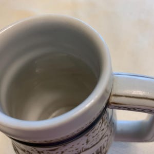 May include: A ceramic beer mug with a white interior and a thick, sturdy handle. The exterior features a textured, off-white surface with dark brown decorative patterns and a prominent dark blue horizontal band encircling the upper section.