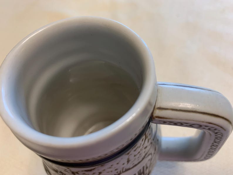 May include: A ceramic beer mug with a white interior and a thick, sturdy handle. The exterior features a textured, off-white surface with dark brown decorative patterns and a prominent dark blue horizontal band encircling the upper section.