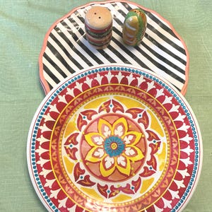 May include: A decorative plate with a red, yellow, and blue floral design. A black and white striped plate with a pink border is behind it. Two ceramic salt and pepper shakers shaped like a hamburger and a pepper are on top.