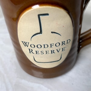 May include: Brown ceramic mug with a handle. The mug features a cream-colored oval with the words "Woodford Reserve" and a bottle outline. The mug is a warm brown color.