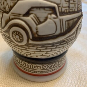 May include: A ceramic beer stein featuring a raised relief design of a vintage car on a cobblestone street. The base includes the text 1936 MG and 1927 Bugatti with a decorative red and black band. The stein has a white finish with brown detailing.